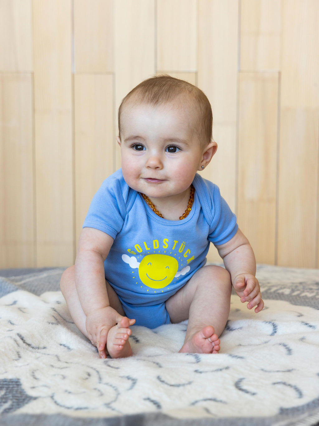 Ein Baby sitzt auf einer Decke und trägt einen hellblauen Baby Body mit "Goldstück"-Print.