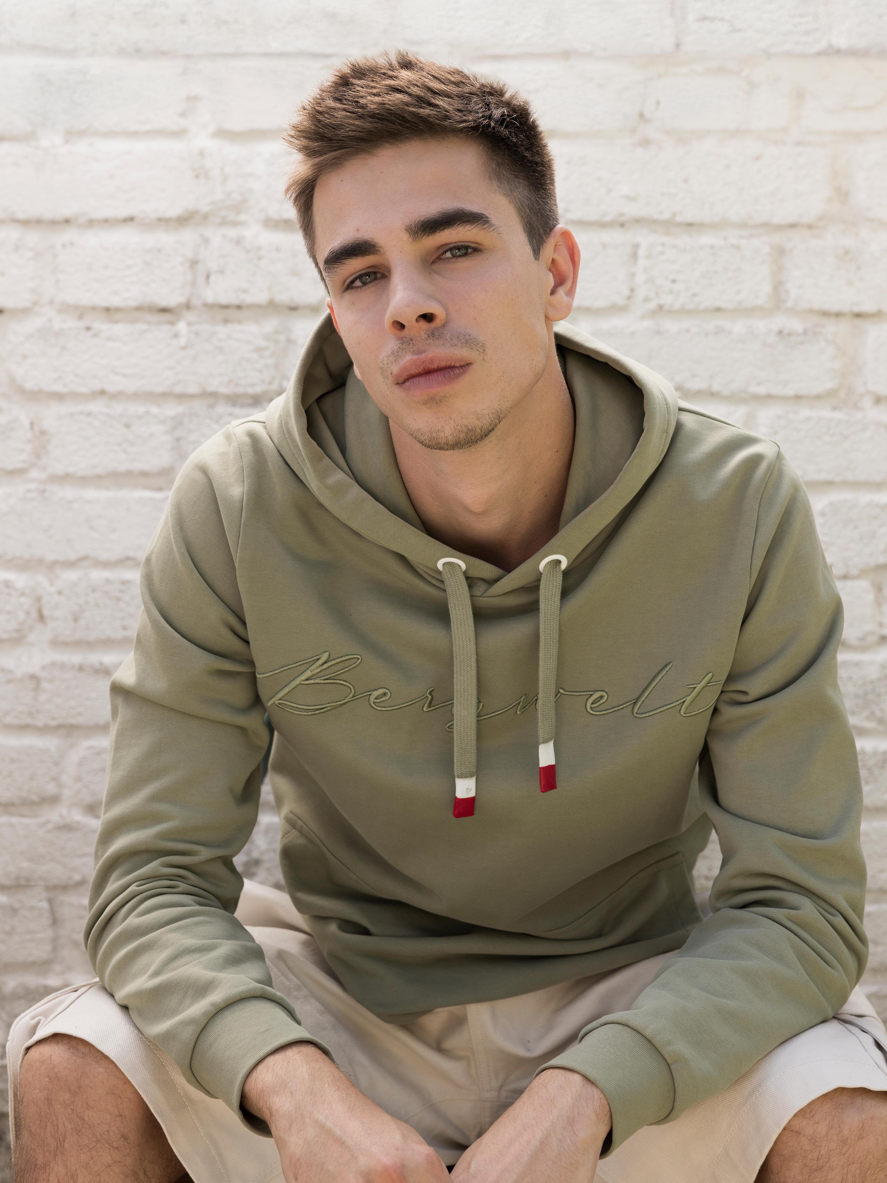 Ein Mann sitzt vor einer hellen Ziegelmauer und trägt den Hoodie "Bergwelt" für Herren in Zementgrau.