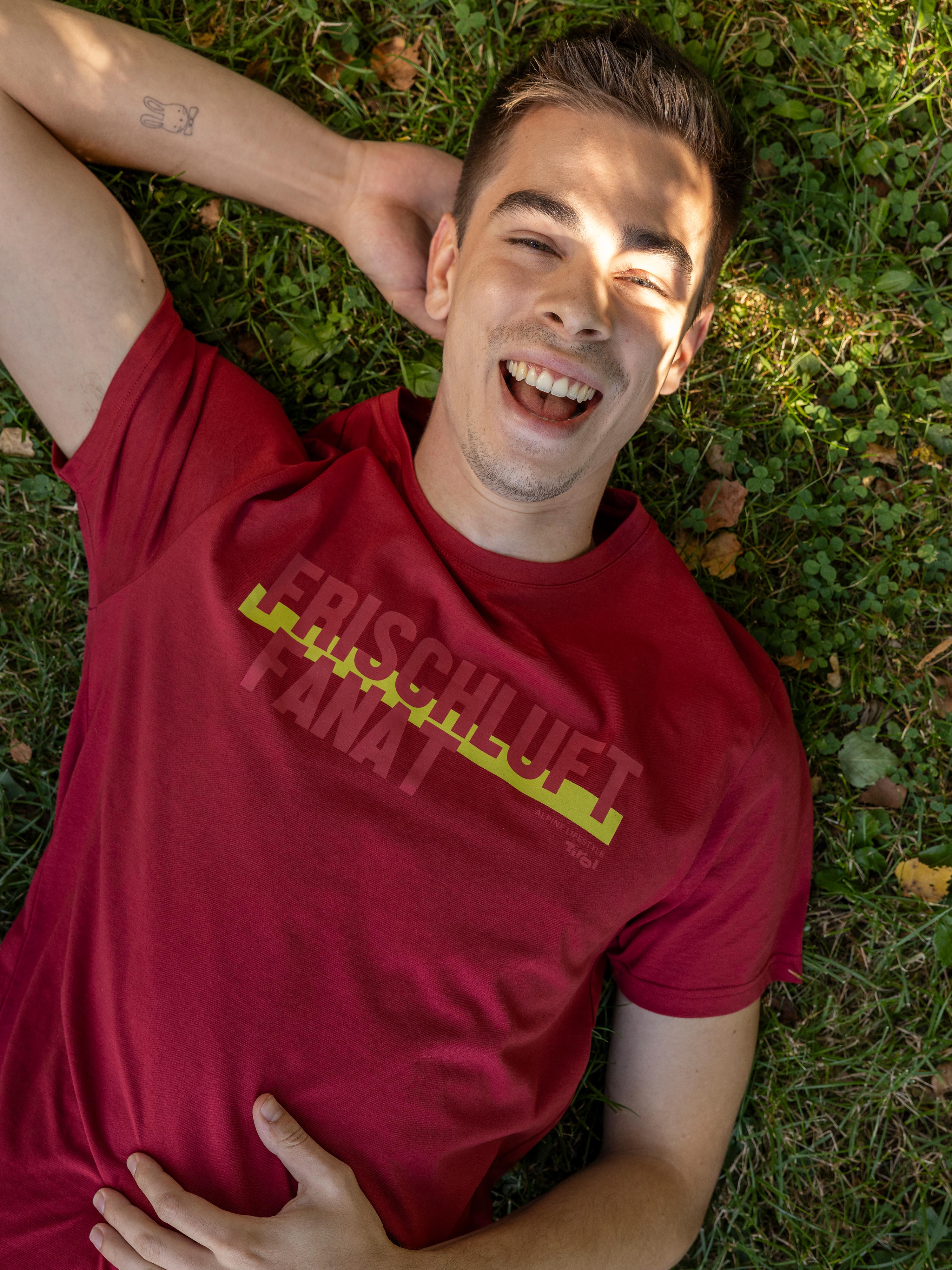 Ein Mann liegt in einer Wiese und trägt das Herren T-Shirt "Frischluft-Fanat" in Rubinrot aus dem Tirol Shop.