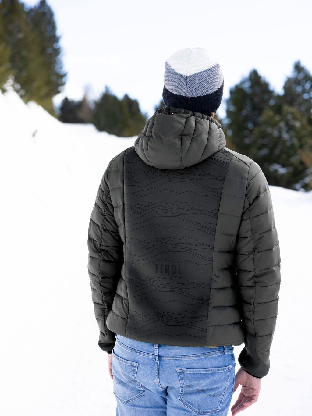 Ein Mann trägt die sportliche, schwarze Hybridjacke "August" aus dem Moments Tirol Shop mit Topographie-3D-Print auf dem Rücken.