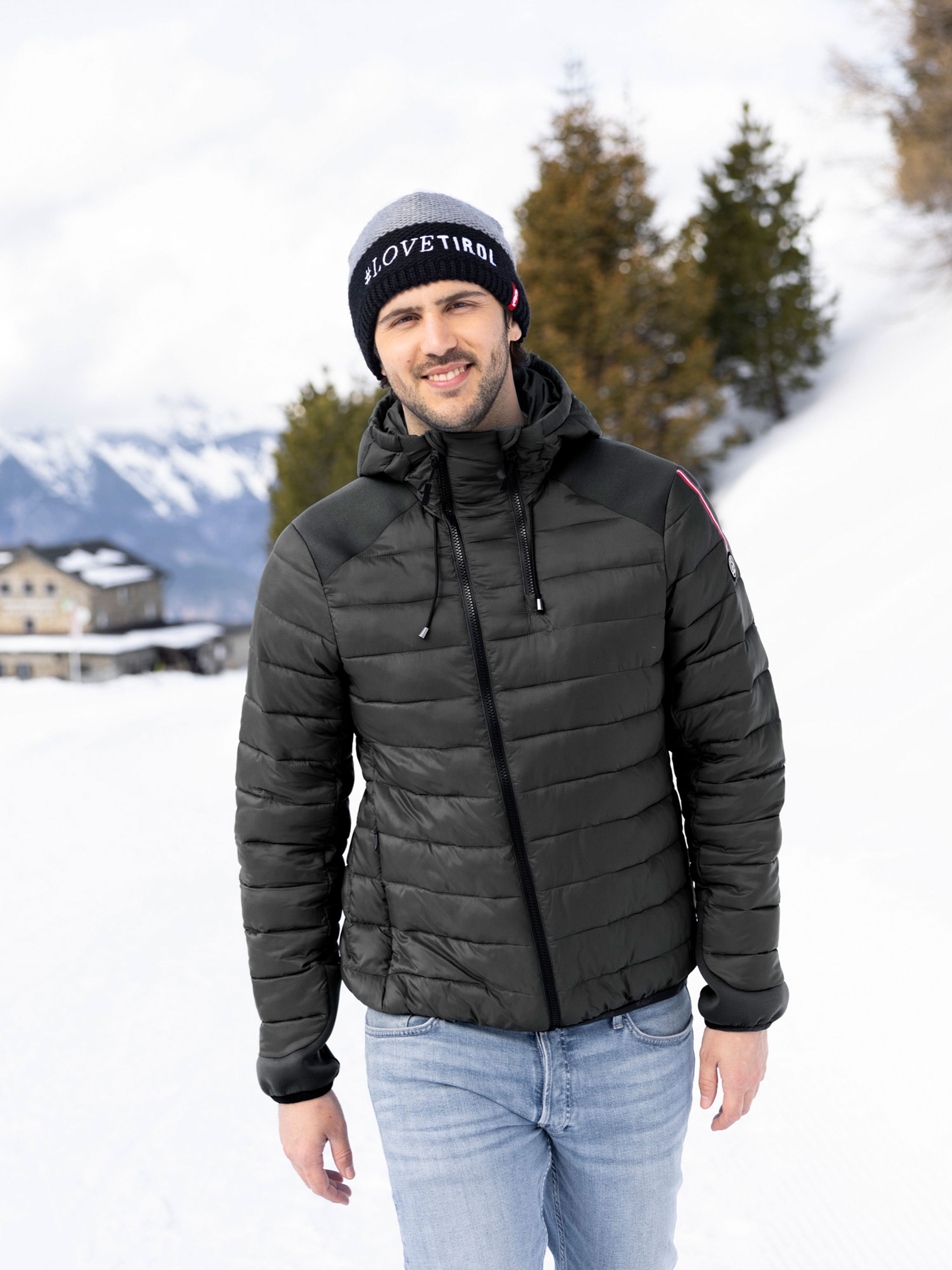 Ein Mann spaziert durch Schnee und trägt die sportliche, schwarze Hybridjacke "August" aus dem Moments Tirol Shop mit asymmetrischem Frontzipper.