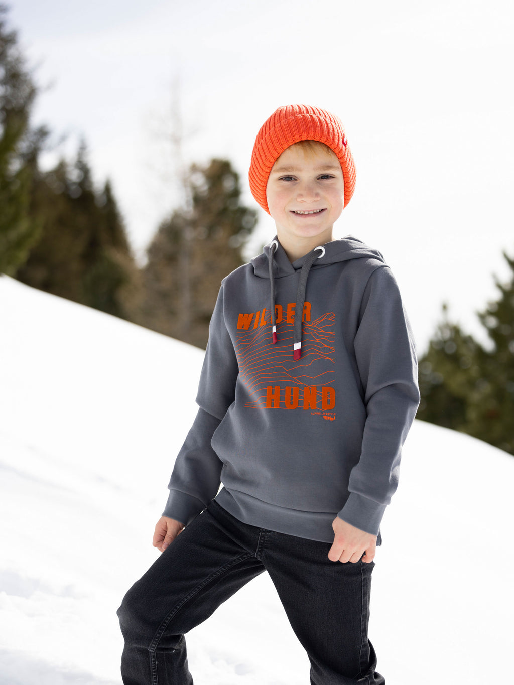 Ein Bub mit oranger Stickmütze namanes "Mika" steht im Schnee und trägt den Kinder Hoodie "Wilder Hund" aus dem Moments Tirol Shop.