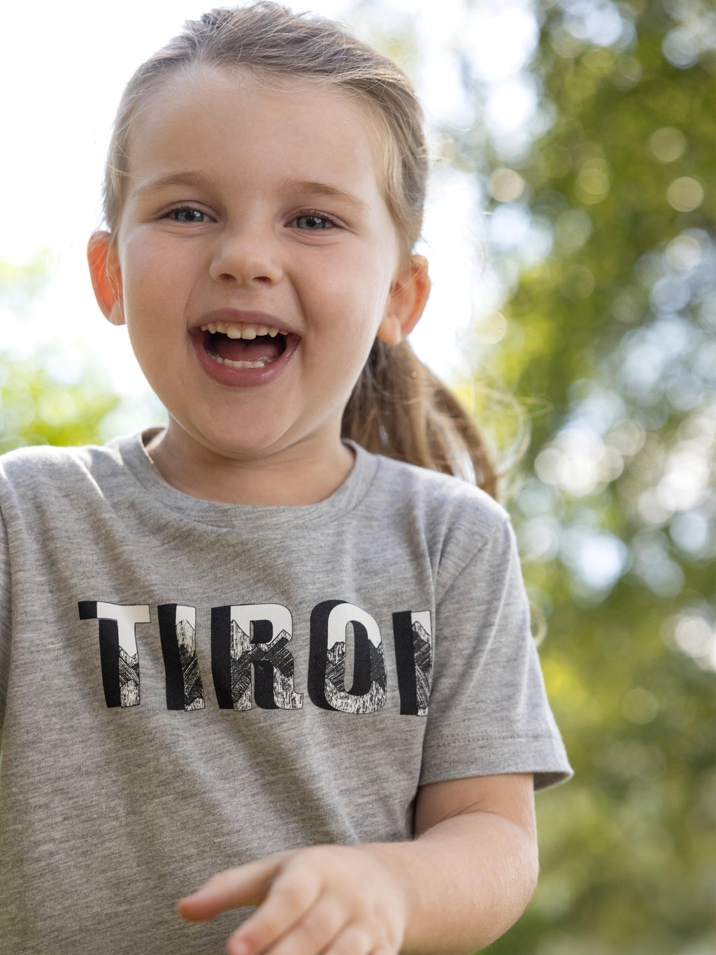 Ein lachendes Mädchen mit langen Haaren trägt das Kinder T-Shirt "Michl" in Grau.