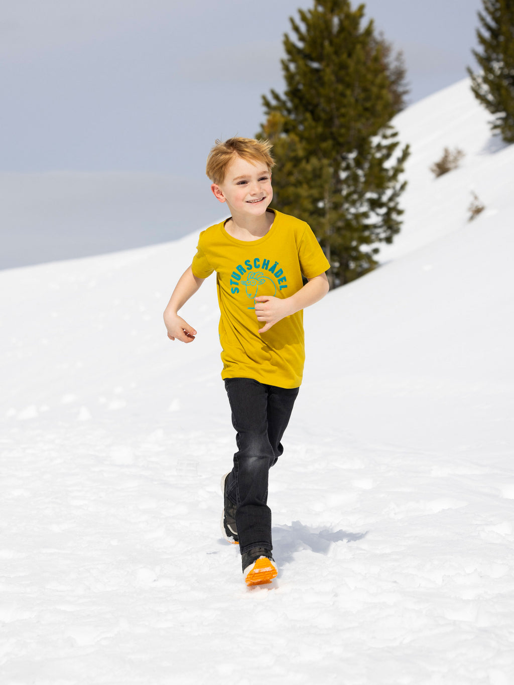 Ein Bub rennt durch den Schnee und trägt das Kinder T-Shirt mit dem "Sturschädel"-Waterbaseprint auf der Brust aus dem Moments Tirol Shop.