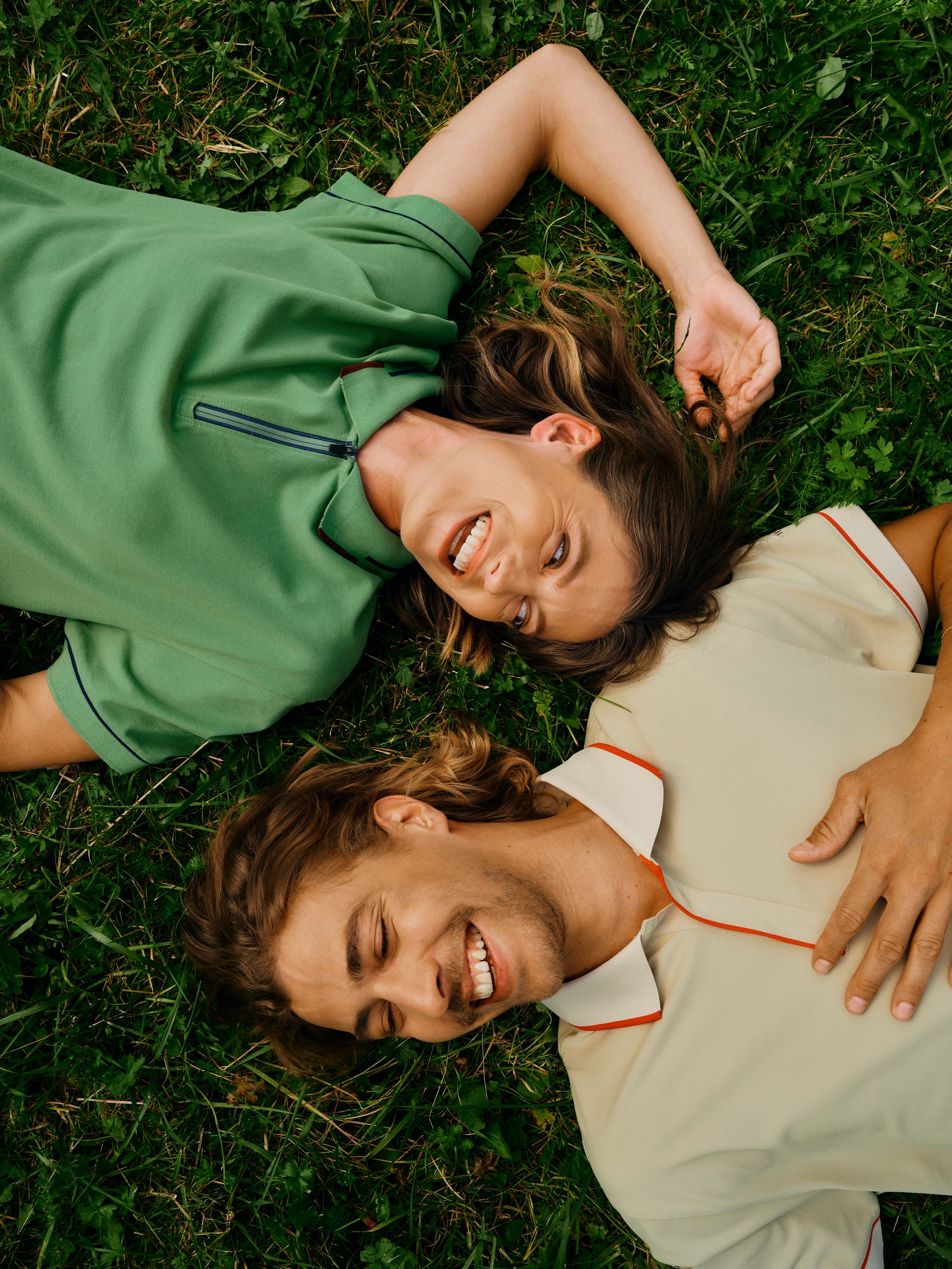 Ein Mann und eine Frau liegen im Gras und tragen das Polo Shirt "Noah" in Grün und das Polo Shirt "Peter-Paul" in Beige.