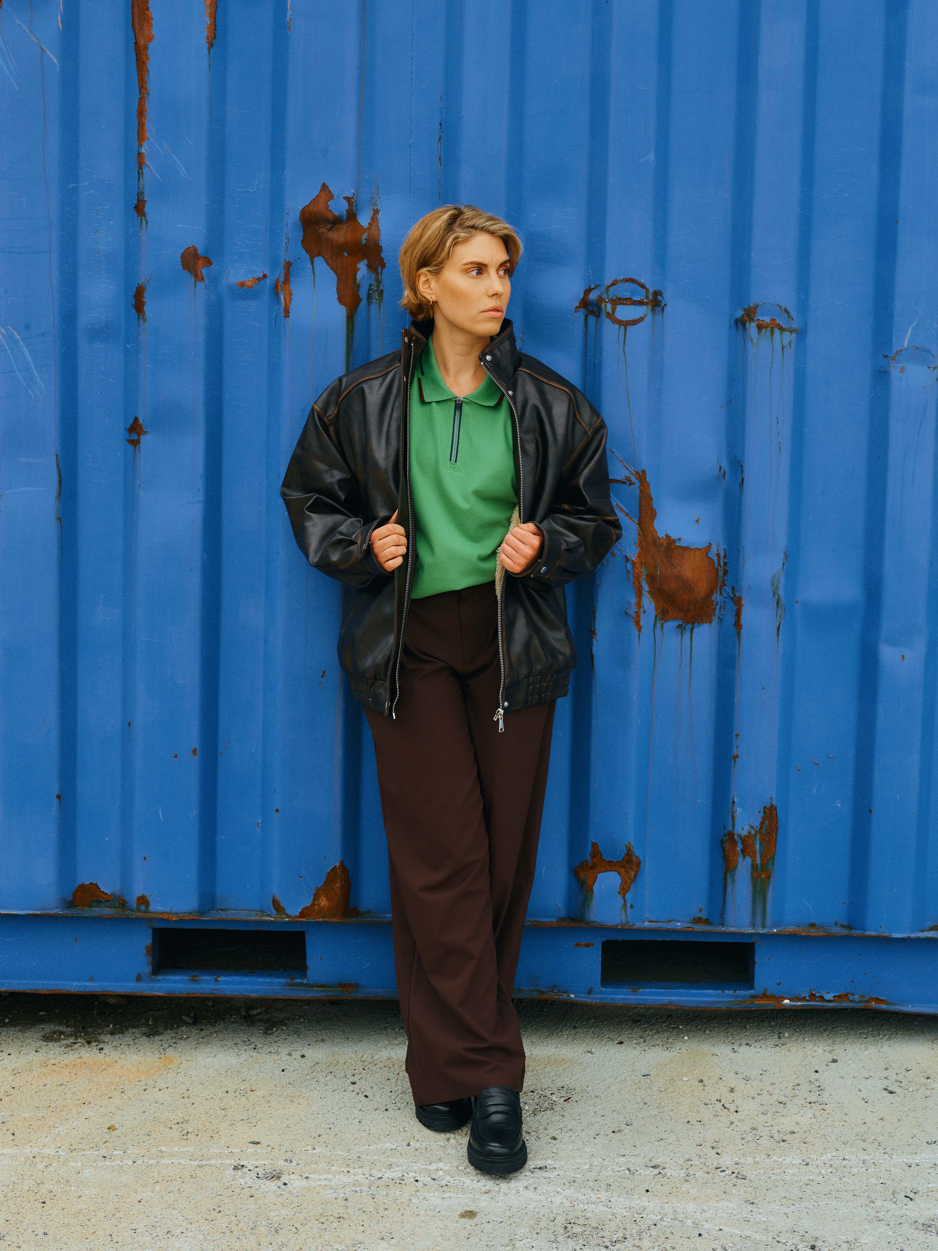 Eine Dame steht vor einem blauen Container und trägt unter einer Lederjacke das Unisex Polo Shirt "Noah" aus dem Tirol Shop.