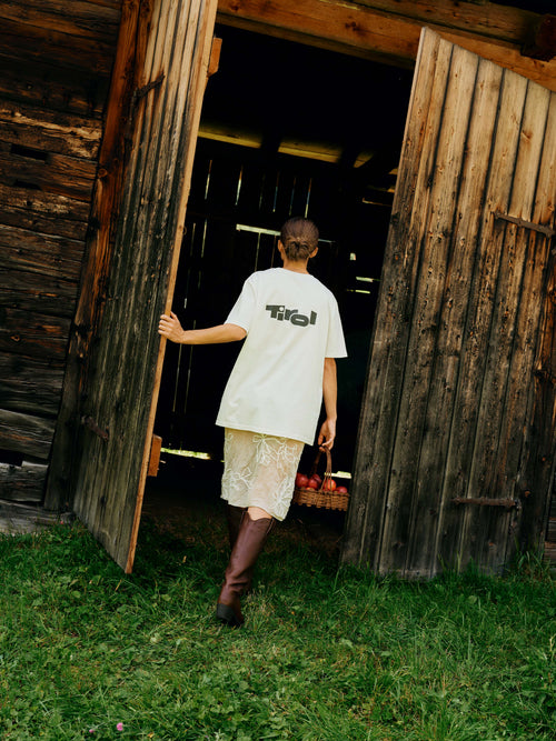 Eine Frau mit braunen Stiefeln und einem Obstkorb in der Hand geht in einen Holzschuppen und trägt das Unisex T-Shirt "Tirol Logo" in Weiß mit großem Tirol Logo Print auf dem Rücken.