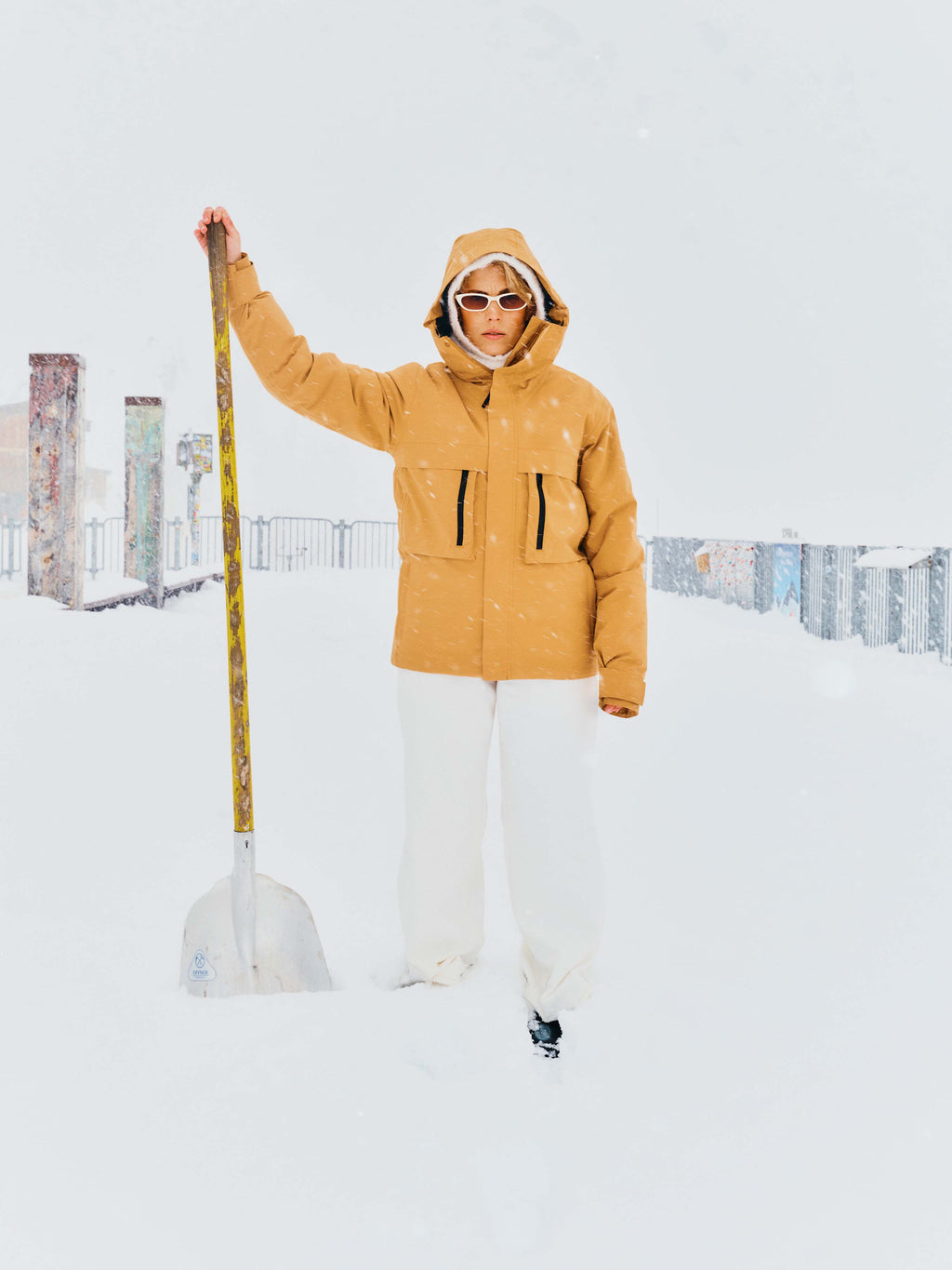 Eine Frau steht mit Schneeschaufel im Schnee, es schneit, sie trägt die Winterjacke "Toni" in Zimt mit Kapuze