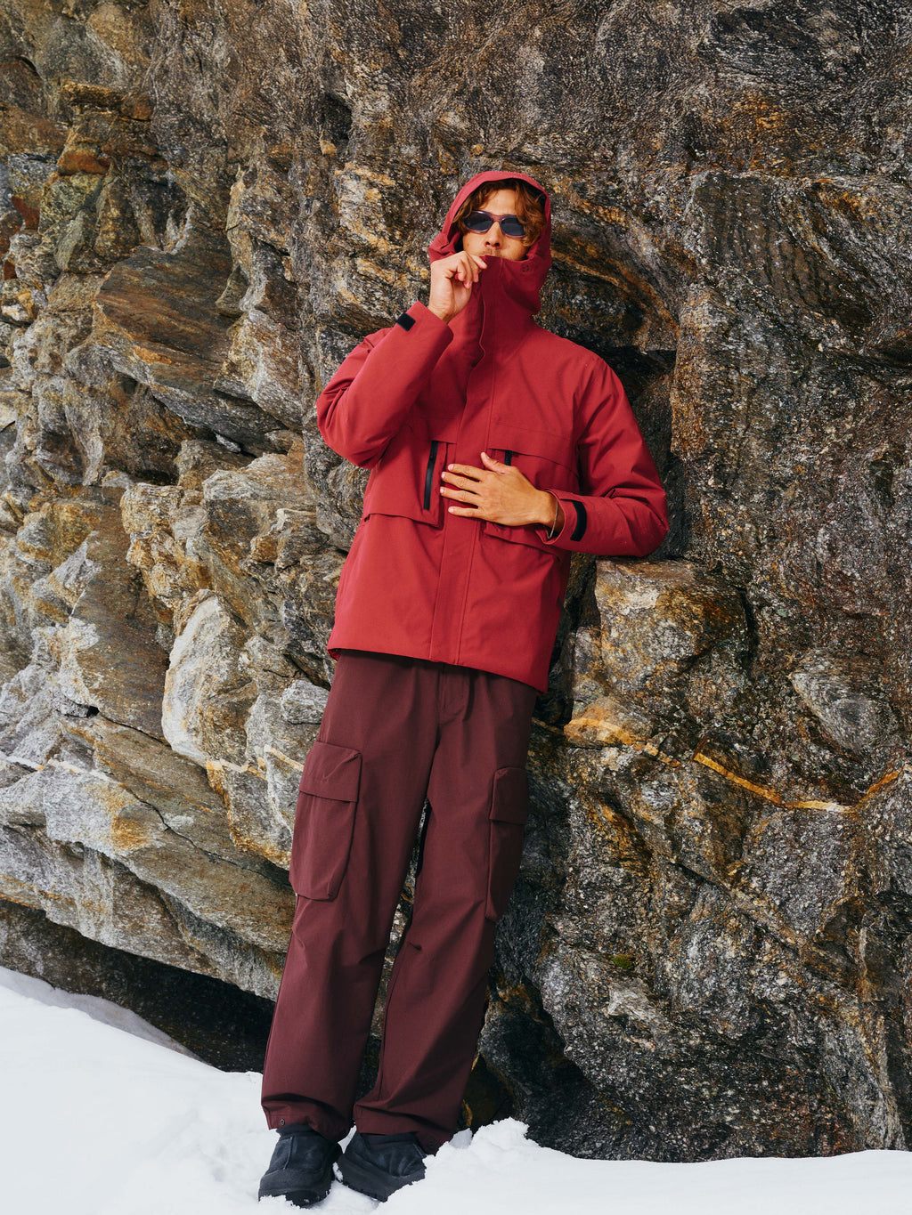 Ein Mann steht vor einem Felsen und trägt die Unisex Winterjacke "Toni" in Rot mit verstellbarer Kapuze