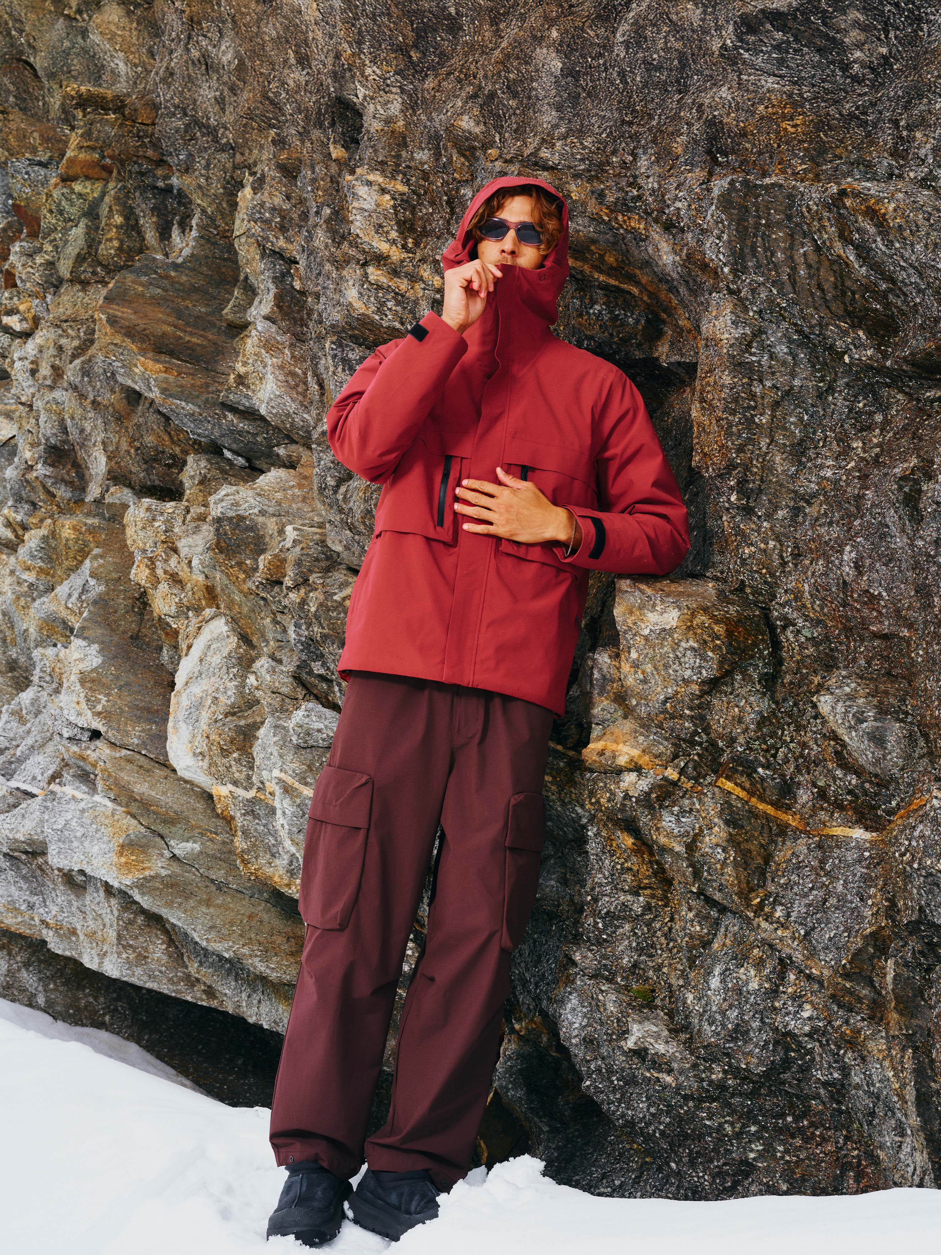 Ein Mann steht vor einem Felsen und trägt die Unisex Winterjacke "Toni" in Rot mit verstellbarer Kapuze